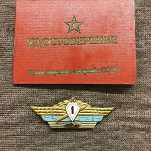 May include: A red booklet with a gold star and the text "УДОСТОВЕРЕНИЕ" in Cyrillic script. Below the booklet is a gold and blue military badge with a red star, the number "1", and a winged spear.