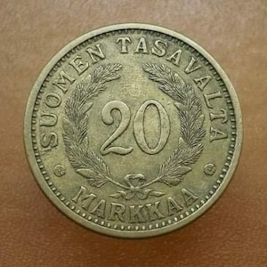 May include: A bronze-colored coin with the text "SUOMEN TASAVALTA" and "20 MARKAA" encircling a central numeral "20" within a wreath. The coin has a detailed edge and is set against a brown background.