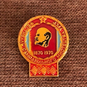 May include: A gold-colored pin with a red and blue design. The pin features a portrait of a man with a beard and the text "1870 1970" and "Республика СССР Самодеятельного Искусства".