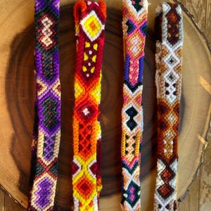 K&ouml;nnte beinhalten: Vier farbenfrohe gewebte Freundschaftsarmb&auml;nder mit geometrischen Mustern. Die Armb&auml;nder weisen eine Vielzahl von Farben auf, darunter Lila, Rot, Orange, Gelb, Rosa und Gr&uuml;n. Jedes Armband hat ein einzigartiges Design und wird auf einer Holzoberfl&auml;che pr&auml;sentiert.