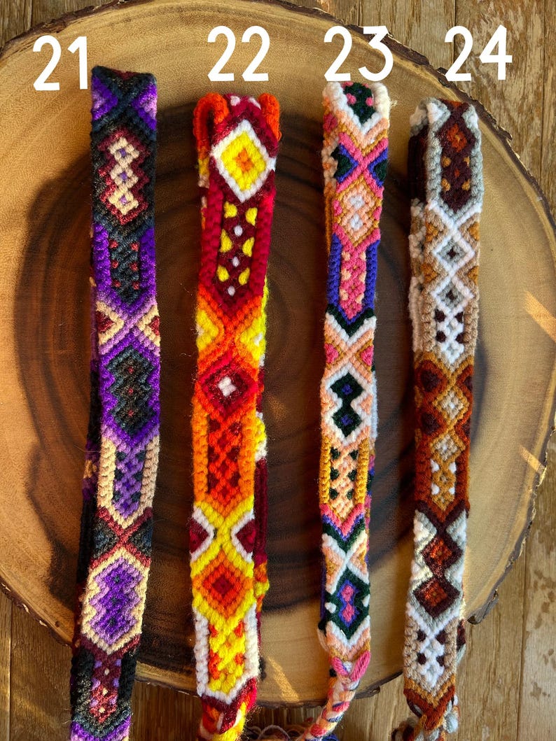 K&ouml;nnte beinhalten: Vier farbenfrohe gewebte Freundschaftsarmb&auml;nder mit geometrischen Mustern. Die Armb&auml;nder weisen eine Vielzahl von Farben auf, darunter Lila, Rot, Orange, Gelb, Rosa und Gr&uuml;n. Jedes Armband hat ein einzigartiges Design und wird auf einer Holzoberfl&auml;che pr&auml;sentiert.