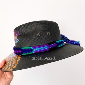 K&ouml;nnte beinhalten: Schwarzer Sonnenhut mit einem gewebten Band in Lila-, Blau- und Gr&uuml;nt&ouml;nen. Der Rand hat ein farbenfrohes Design mit dem Text "Solei Azul". Der Hut hat eine breite Krempe und eine dekorative Quaste.