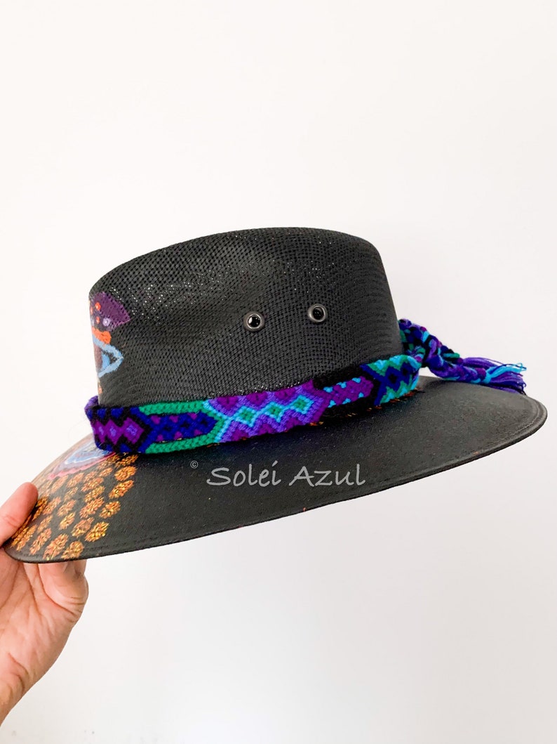 K&ouml;nnte beinhalten: Schwarzer Sonnenhut mit einem gewebten Band in Lila-, Blau- und Gr&uuml;nt&ouml;nen. Der Rand hat ein farbenfrohes Design mit dem Text "Solei Azul". Der Hut hat eine breite Krempe und eine dekorative Quaste.