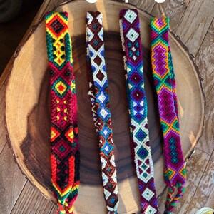 K&ouml;nnte beinhalten: Vier farbenfrohe gewebte Freundschaftsarmb&auml;nder mit geometrischen Mustern. Die Armb&auml;nder weisen eine Vielzahl von Farben auf, darunter Rot, Gr&uuml;n, Gelb, Blau und Lila. Jedes Armband hat ein einzigartiges Design und wird auf einer Holzoberfl&auml;che pr&auml;sentiert.