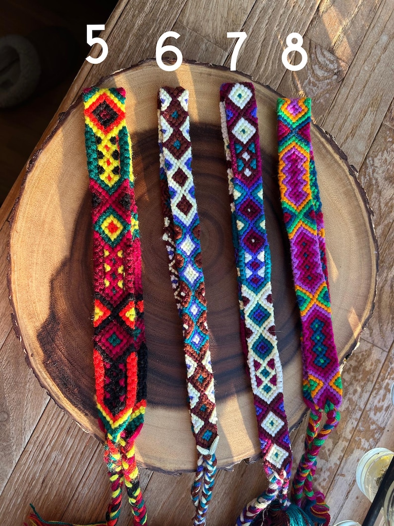 K&ouml;nnte beinhalten: Vier farbenfrohe gewebte Freundschaftsarmb&auml;nder mit geometrischen Mustern. Die Armb&auml;nder weisen eine Vielzahl von Farben auf, darunter Rot, Gr&uuml;n, Gelb, Blau und Lila. Jedes Armband hat ein einzigartiges Design und wird auf einer Holzoberfl&auml;che pr&auml;sentiert.