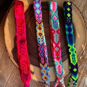 K&ouml;nnte beinhalten: Vier farbenfrohe Freundschaftsarmb&auml;nder mit aufwendigen Webmustern. Die Armb&auml;nder weisen leuchtende Farbt&ouml;ne von Rot, Blau, Rosa, Lila und Gr&uuml;n auf. Jedes Armband hat ein einzigartiges geometrisches Design und eine Quaste am Ende. Die Armb&auml;nder werden auf einer Holzoberfl&auml;che pr&auml;sentiert.