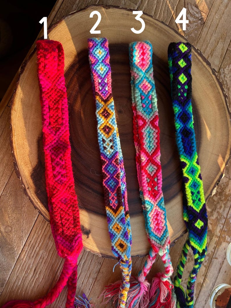 K&ouml;nnte beinhalten: Vier farbenfrohe Freundschaftsarmb&auml;nder mit aufwendigen Webmustern. Die Armb&auml;nder weisen leuchtende Farbt&ouml;ne von Rot, Blau, Rosa, Lila und Gr&uuml;n auf. Jedes Armband hat ein einzigartiges geometrisches Design und eine Quaste am Ende. Die Armb&auml;nder werden auf einer Holzoberfl&auml;che pr&auml;sentiert.