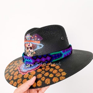 K&ouml;nnte beinhalten: Schwarzer Hut mit breiter Krempe, verziert mit einem farbenfrohen Totenkopf-Design und floralen Akzenten. Der Hut hat ein gewebtes Band in Lila, Blau und Gr&uuml;n. Der Rand ist mit orangefarbenen Blumenmustern verziert.