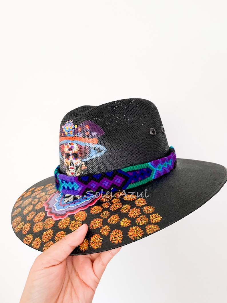 K&ouml;nnte beinhalten: Schwarzer Hut mit breiter Krempe, verziert mit einem farbenfrohen Totenkopf-Design und floralen Akzenten. Der Hut hat ein gewebtes Band in Lila, Blau und Gr&uuml;n. Der Rand ist mit orangefarbenen Blumenmustern verziert.