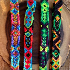 K&ouml;nnte beinhalten: Vier farbenfrohe gewebte Freundschaftsarmb&auml;nder mit geometrischen Mustern. Die Armb&auml;nder weisen lebendige Farbt&ouml;ne von Rosa, Blau, Gr&uuml;n, Rot und Gelb auf. Jedes Armband hat ein einzigartiges Design, das komplizierte Kn&uuml;pftechniken zeigt. Die Armb&auml;nder werden auf einer Holzoberfl&auml;che pr&auml;sentiert.