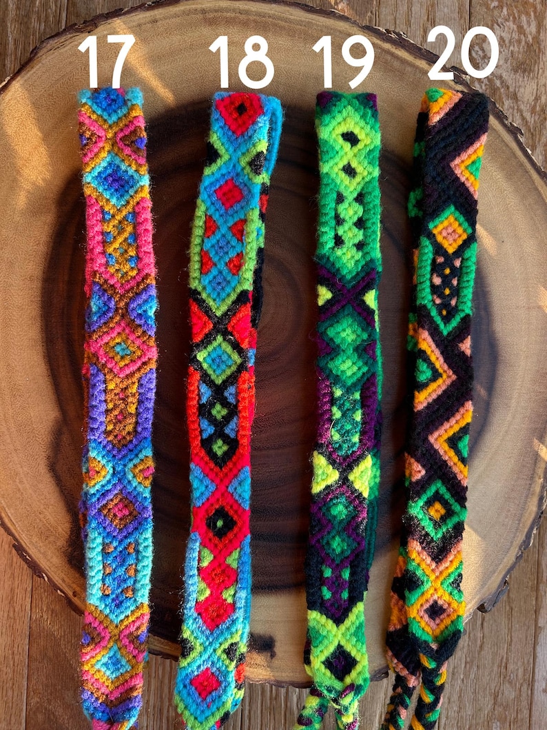 K&ouml;nnte beinhalten: Vier farbenfrohe gewebte Freundschaftsarmb&auml;nder mit geometrischen Mustern. Die Armb&auml;nder weisen lebendige Farbt&ouml;ne von Rosa, Blau, Gr&uuml;n, Rot und Gelb auf. Jedes Armband hat ein einzigartiges Design, das komplizierte Kn&uuml;pftechniken zeigt. Die Armb&auml;nder werden auf einer Holzoberfl&auml;che pr&auml;sentiert.