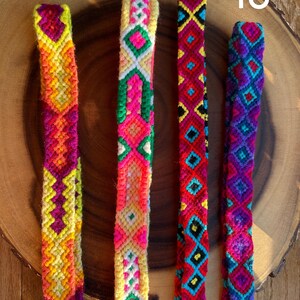K&ouml;nnte beinhalten: Vier farbenfrohe gewebte Freundschaftsarmb&auml;nder mit geometrischen Mustern. Die Armb&auml;nder weisen lebendige Farbt&ouml;ne von Gelb, Orange, Rosa, Rot, Blau und Lila auf. Jedes Armband hat ein einzigartiges Design, das komplizierte Kn&uuml;pftechniken zeigt. Die Armb&auml;nder werden auf einer Holzoberfl&auml;che pr&auml;sentiert.