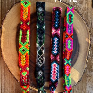 K&ouml;nnte beinhalten: Vier farbenfrohe gewebte Freundschaftsarmb&auml;nder mit komplizierten geometrischen Mustern. Die Armb&auml;nder weisen leuchtende Farbt&ouml;ne von Rot, Orange, Gr&uuml;n, Pink, Blau und Schwarz auf. Jedes Armband wird auf einer Holzscheibe mit den Nummern 9, 10, 11 und 12 dar&uuml;ber pr&auml;sentiert.