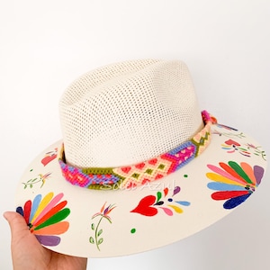 K&ouml;nnte beinhalten: Ein cremefarbener Strohhut mit einem farbenfrohen gewebten Band und bemalten Blumenmustern. Der Rand weist handgemalte Blumen in verschiedenen Farben auf, darunter Rot, Orange, Gelb und Lila. Der Hut wird von einer Hand gehalten.