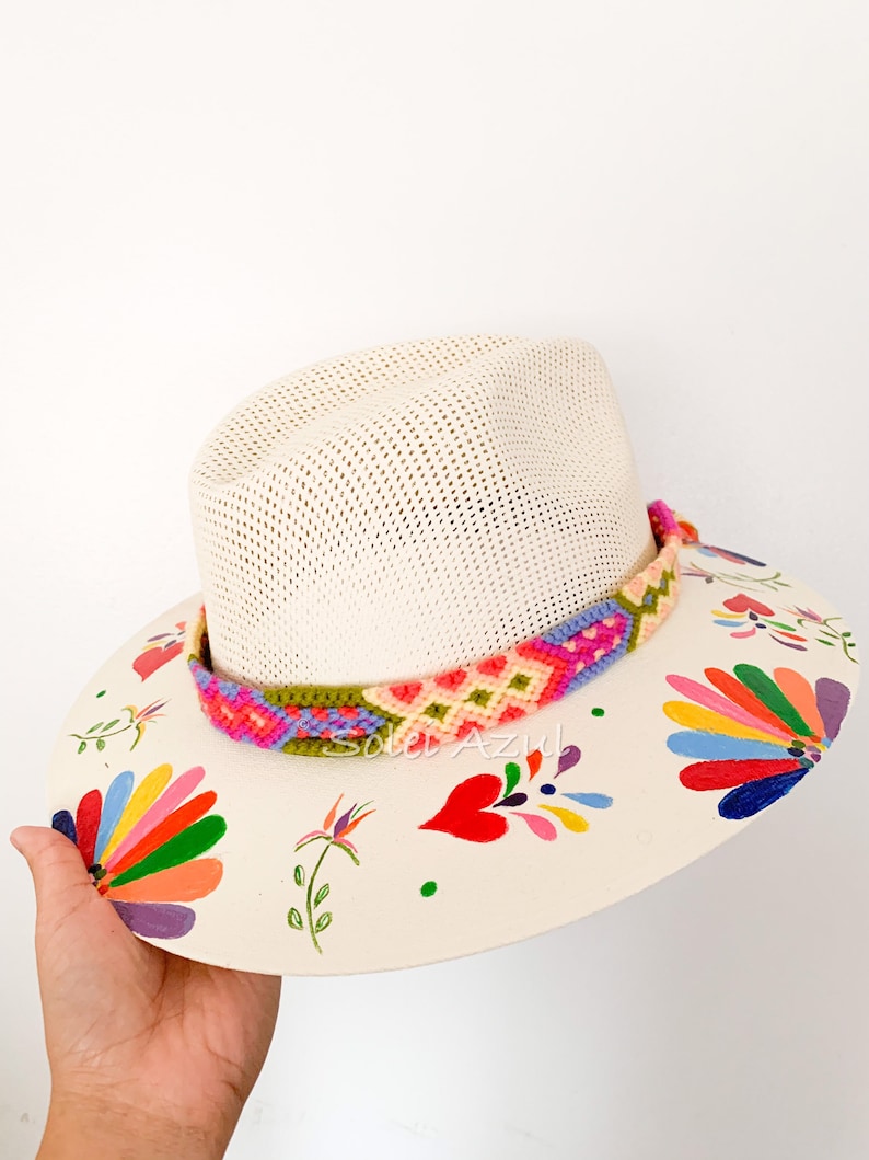 K&ouml;nnte beinhalten: Ein cremefarbener Strohhut mit einem farbenfrohen gewebten Band und bemalten Blumenmustern. Der Rand weist handgemalte Blumen in verschiedenen Farben auf, darunter Rot, Orange, Gelb und Lila. Der Hut wird von einer Hand gehalten.