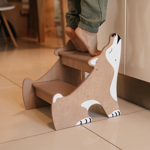 May include: A wooden two-step stool shaped like a dog with white and black accents. The stool is made of light brown wood and has a white painted face with a black nose and eyes. The stool is standing on a light brown tiled floor.