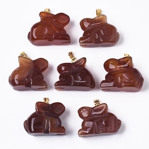 May include: Seven brown rabbit pendants with gold-colored bails. The pendants are carved from a translucent, reddish-brown stone. Each rabbit is depicted in a seated position with its ears perked up.
