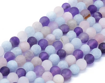 May include: Close-up of strands of round gemstone beads in various shades of purple, lavender, light blue, and white. The beads have a matte finish and are arranged closely together, creating a textured surface. The beads are likely used for jewelry making.