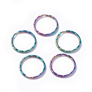 May include: Five iridescent key rings with a hammered texture are arranged on a white background. The rings display a spectrum of colors, including blues, greens, and purples, creating a vibrant and eye-catching effect. These key rings are suitable for various uses.