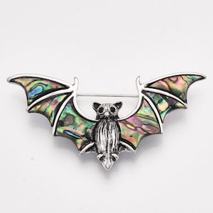 May include: A silver-toned bat brooch with iridescent wings. The bat's wings display a vibrant array of colors, including green, blue, and purple. The brooch has a pin closure on the back and is set against a white background.