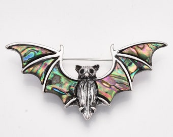 May include: A silver-toned bat brooch with iridescent wings. The bat's wings display a vibrant array of colors, including green, blue, and purple. The brooch has a pin closure on the back and is set against a white background.