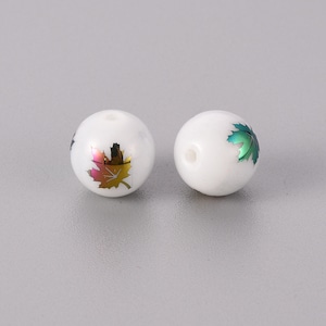 May include: Two white, round beads with a maple leaf design. One leaf is a mix of gold, orange, and pink, while the other is green. Each bead has a small hole for stringing.