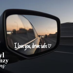 May include: A car side mirror reflecting a highway scene at dusk. White text in the mirror reads "I love you, drive safe" with a heart symbol. The words "Vinyl Frenzy Creations" are at the bottom.
