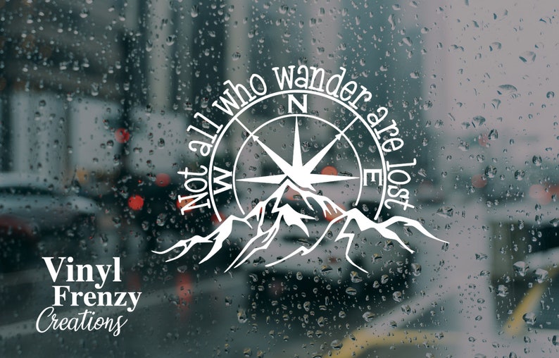 May include: A white vinyl decal featuring a compass rose and mountain range graphic with the text "Not all who wander are lost" arranged in a circular pattern, accompanied by the text "Vinyl Frenzy Creations" in the bottom left corner.