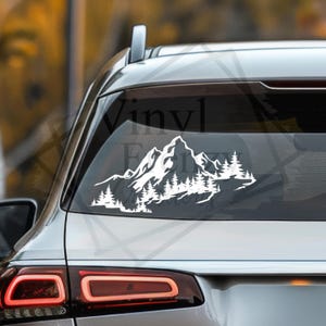 Op de afbeelding: Een witte vinyl sticker aangebracht op de achterruit van een voertuig, met een gedetailleerde afbeelding van een bergketen met grillige toppen en een dicht dennenbos aan de voet, wat een natuur-geïnspireerd landschapsontwerp creëert.