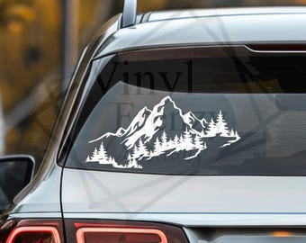 May include: A white vinyl decal applied to the rear window of a vehicle, featuring a detailed graphic of a mountain range with jagged peaks and a dense forest of pine trees at the base, creating a nature-inspired landscape design.