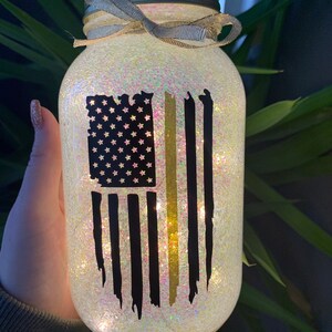 Könnte beinhalten: Ein klares Glasgefäß mit goldenem Deckel und weißem Band. Das Gefäß ist mit schimmerndem Glitzer gefüllt und zeigt einen schwarzen und goldenen amerikanischen Flaggenaufkleber mit einem dünnen gelben Streifen.