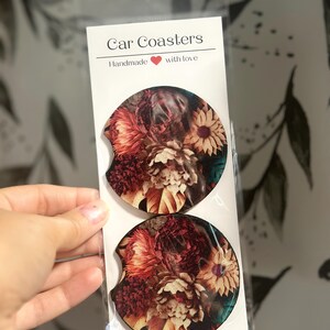 May include: Two round car coasters with a floral design. The coasters feature a variety of flowers in shades of red, orange, yellow, and white. The text "Car Coasters" and "Handmade with love" is printed on the white packaging.