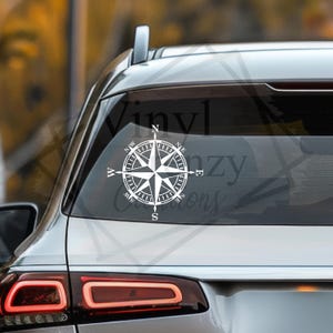 May include: A white vinyl decal of a nautical compass rose featuring cardinal directions N, S, E, and W, along with intermediate directional markers, applied to the center of a vehicle's rear window, displaying a clean and sharp graphic design.