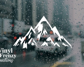 May include: White vinyl decal of a mountain range silhouette on a blurry background of a rainy city street. The decal is for a car window or other smooth surface. The text "Vinyl Frenzy Creations" is visible at the bottom of the image.
