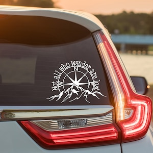 May include: A white vinyl decal applied to a car rear window featuring a compass rose design centered above a mountain range silhouette with the text "Not all who wander are lost" curved along the top edge of the compass graphic.