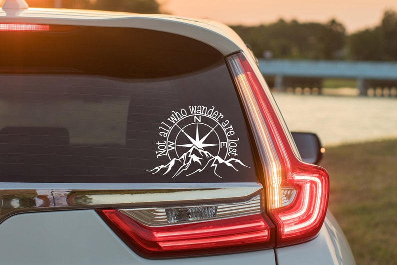 May include: A white vinyl decal applied to a car rear window featuring a compass rose design centered above a mountain range silhouette with the text "Not all who wander are lost" curved along the top edge of the compass graphic.