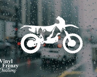 May include: A white vinyl decal of a dirt bike silhouette featuring a negative space mountain and pine tree forest design within the frame. The text Vinyl Frenzy Creations is printed in white in the lower left corner of the image.