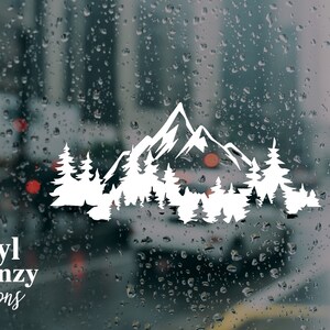 May include: A white vinyl decal featuring a stylized mountain range silhouette layered behind a dense forest of pine trees. The design is applied to a glass surface covered in raindrops, with the text Vinyl Frenzy Creations printed in white.
