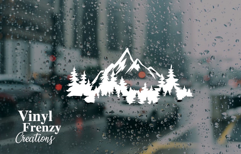May include: A white vinyl decal featuring a stylized mountain range silhouette layered behind a dense forest of pine trees. The design is applied to a glass surface covered in raindrops, with the text Vinyl Frenzy Creations printed in white.