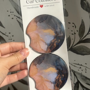 May include: Two round car coasters with an abstract design featuring shades of blue, gray, pink, and gold. The coasters are packaged in a clear plastic bag with a white label that reads "Car Coasters Handmade with love".
