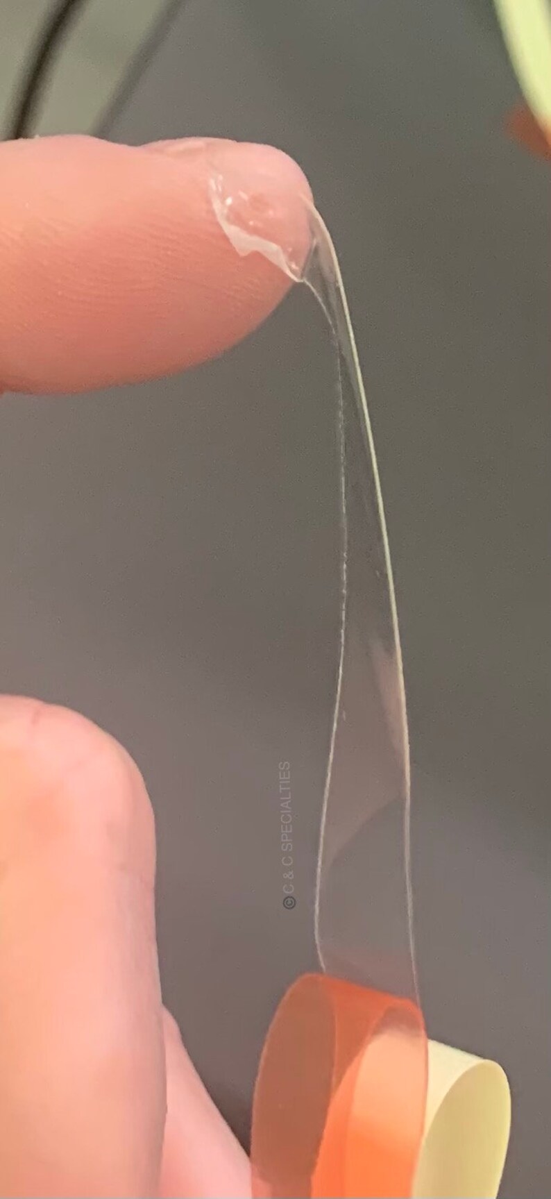 May include: A close-up of clear, transparent tape being stretched and pulled apart. The tape is thin and flexible, and it is being held by a finger. The tape is being pulled apart to show its elasticity and strength. &copy; C&C Specialties