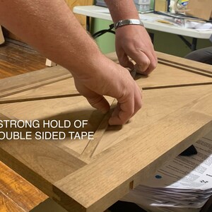 May include: A close-up of a wooden door with a geometric design. A person is using double-sided tape to secure the door. The text "STRONG HOLD OF DOUBLE SIDED TAPE" is visible in the image.