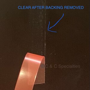 May include: A close-up of a clear adhesive tape with a pink backing. The backing has been partially removed, revealing the clear adhesive. The text "CLEAR AFTER BACKING REMOVED" is visible in the top right corner of the image.