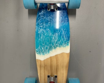 Könnte beinhalten: Ein Skateboard aus Holz mit blauer und weißer Kunstharz-Kunst, die Ozeanwellen darstellt. Das Board hat blaue Räder und silberne Achsen.