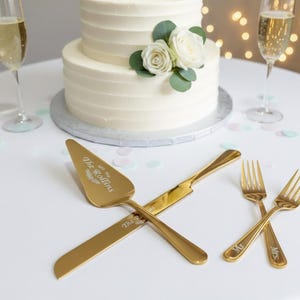 以下が含まれることがあります： 花飾りのついた2段の白いウェディングケーキ、シャンパングラス、金色のサービング用具。ケーキサーバーとナイフは交差しており、フォークが近くにあります。ケーキサーバーには「The Rollins」の文字が刻まれています。