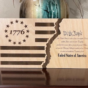 Könnte beinhalten: Ein Holzschild mit einem abgenutzten Design der amerikanischen Flagge. Die Flagge ist in zwei Hälften geteilt, wobei die obere Hälfte die Sterne und Streifen zeigt und die untere Hälfte den Text "We the People" aus der US-Verfassung. Das Jahr "1776" ist in der Mitte der Sterne eingraviert.