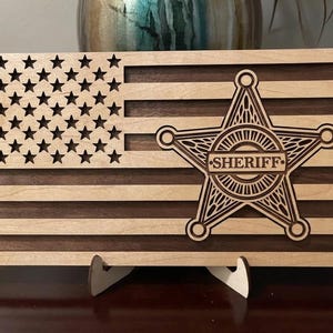 May include: A rectangular wooden wall art piece featuring a laser-cut American flag design with alternating light and dark wood stripes. A prominent five-pointed sheriff star badge with the word SHERIFF engraved in the center is layered on top.