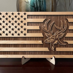 Può includere: Una bandiera americana in legno con un ritaglio a forma di testa di cervo al centro. La bandiera è realizzata in legno di acero e noce ed è dotata di un supporto.