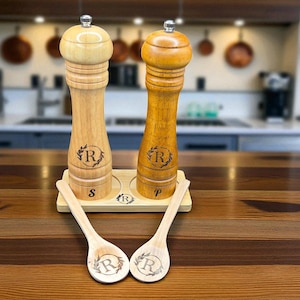 Puede incluir: Molinillos de sal y pimienta de madera con cucharas de madera a juego. Los molinillos son de madera clara y oscura, grabados con una letra y una corona de laurel. Las cucharas también tienen el mismo grabado. El juego se encuentra en una bandeja de madera.