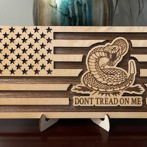 May include: A wooden American flag with a rattlesnake and the words "Don't Tread On Me" engraved on it. The flag is mounted on a small wooden stand.